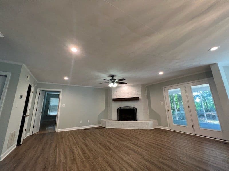 Spacious living room with wood floors, ceiling fan, and fireplace, featuring natural light from doors.