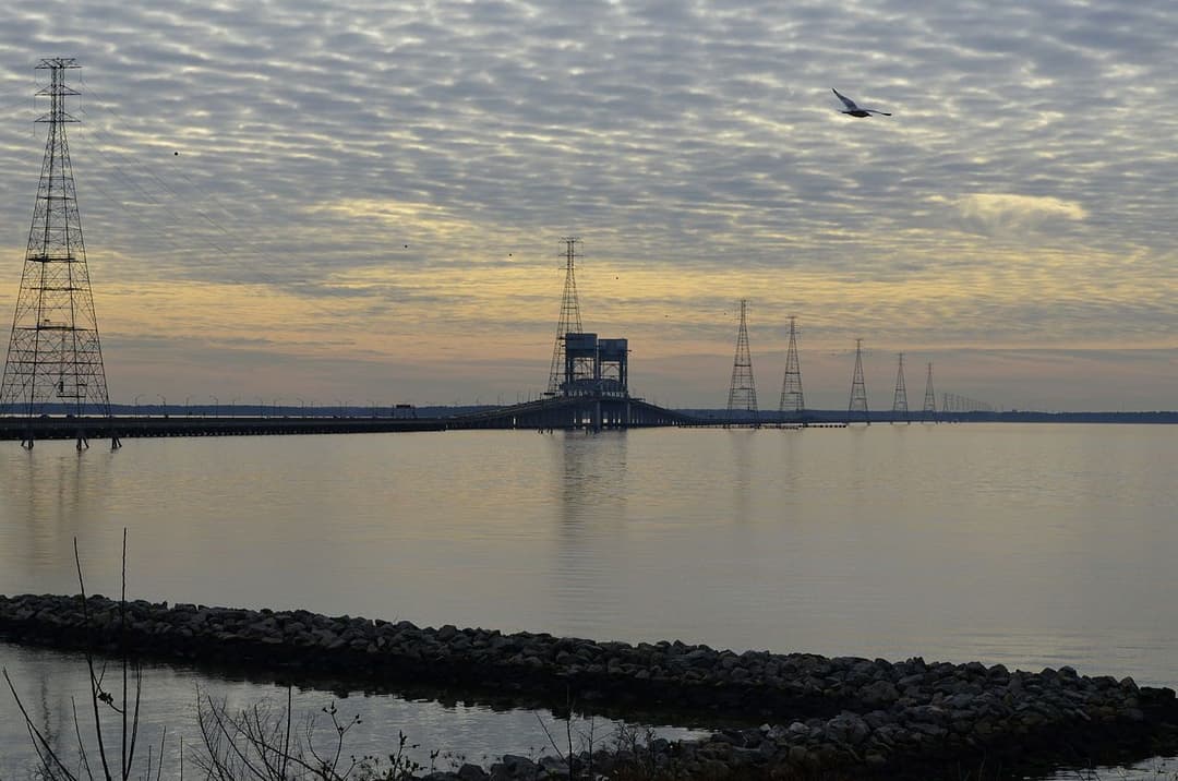 Bridge over calm water at sunset with power lines and a cloudy sky.